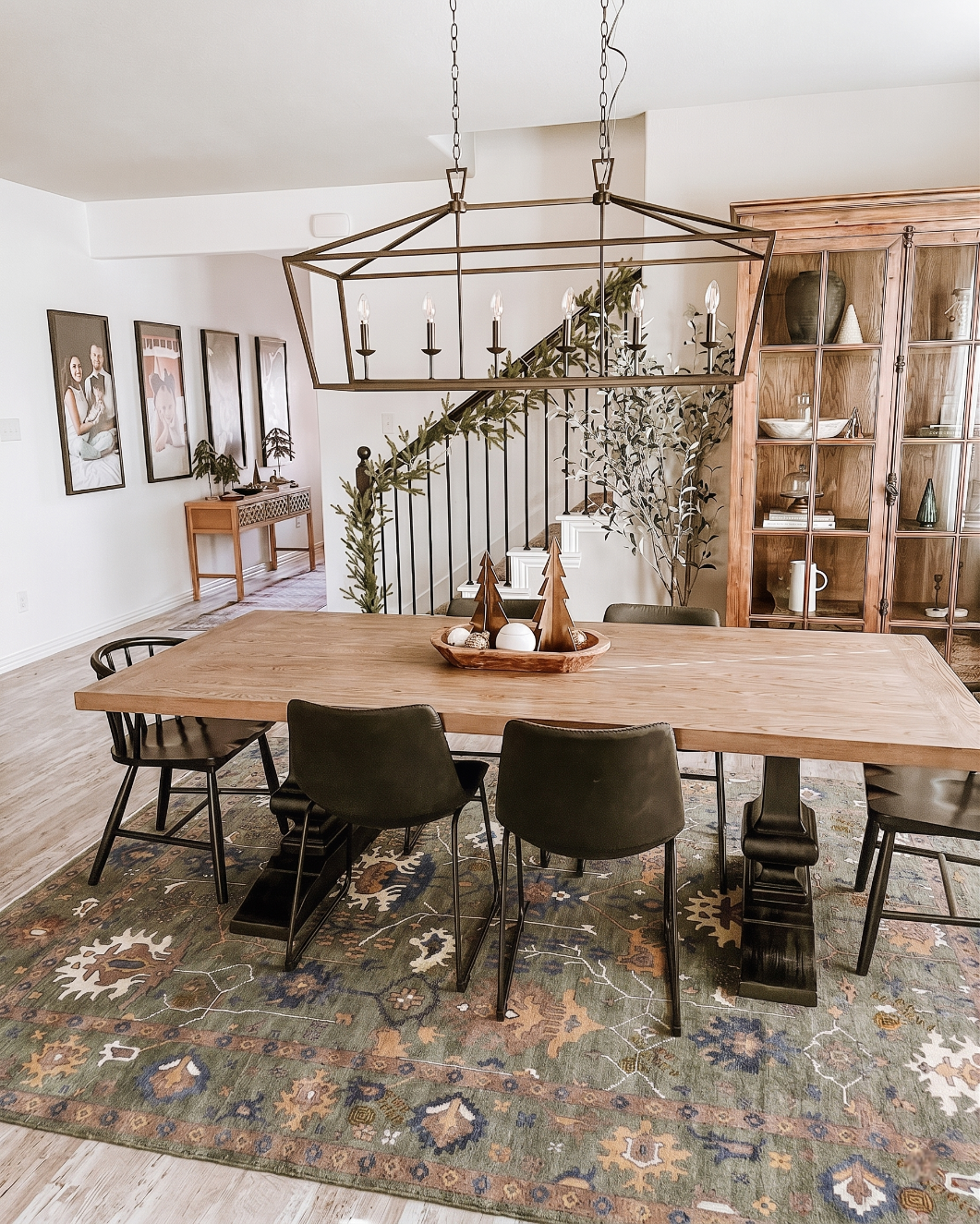 A rustic wooden dining table with black chairs in a styled dining room, featuring a large patterned area rug, a geometric metal pendant light, and a wooden hutch in the background.